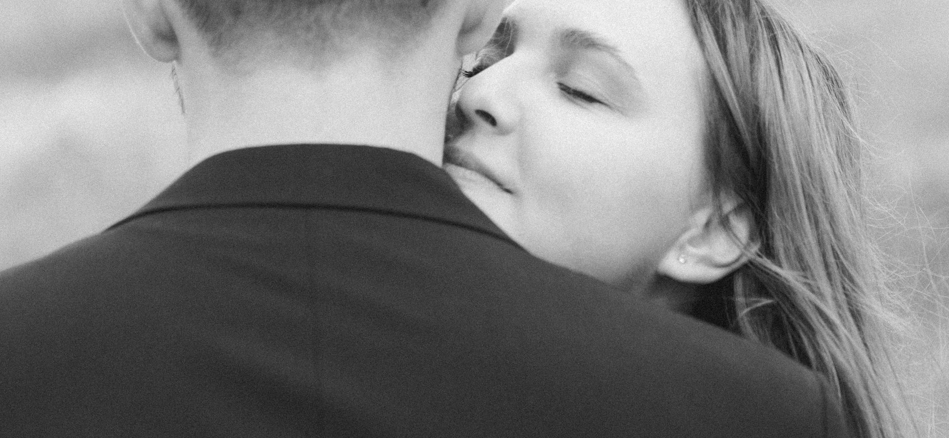 Black and white photography of a woman leaning in to kiss her groom on the neck.