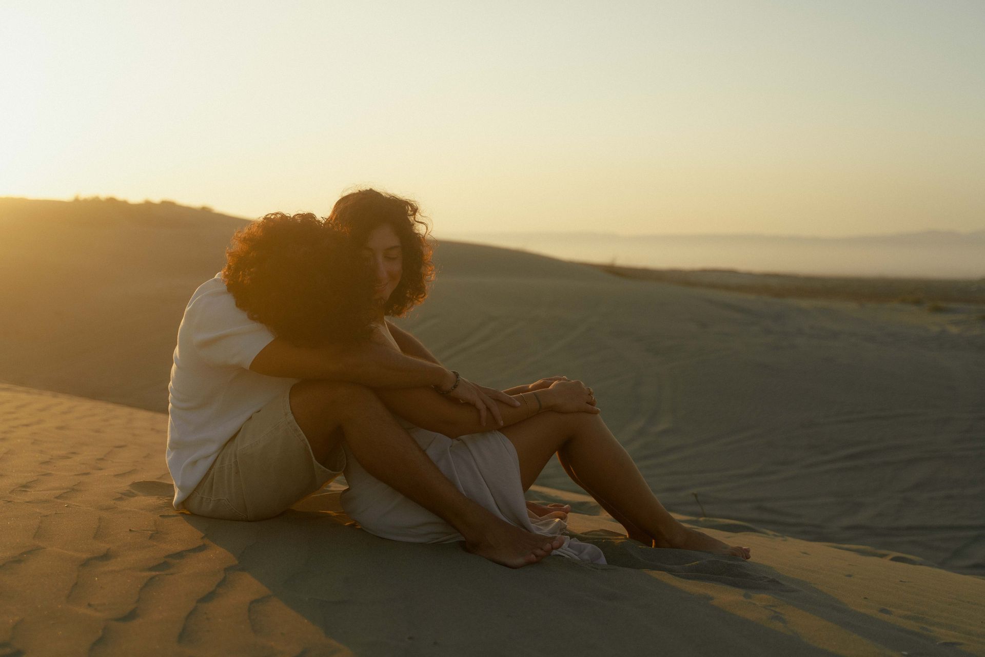 A couple sitting on a sand dune, hugging and cuddling at sunset.