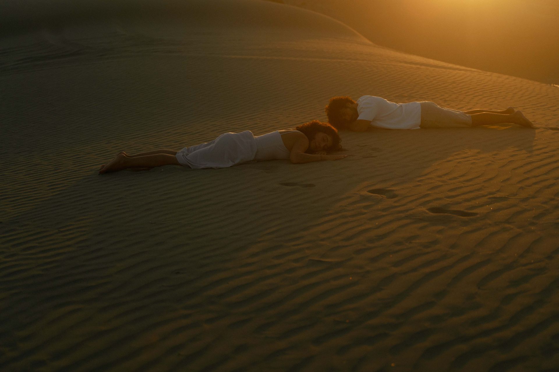 Two people laying on a sand dune at sunset, facing each other.