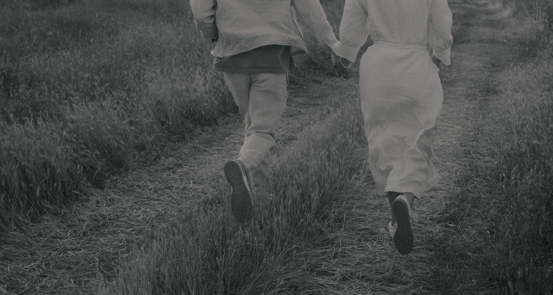 Black and white photography of a man and a woman running through a field, hand in hand in wedding clothes