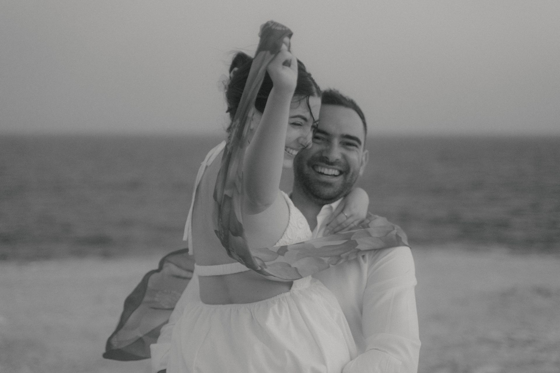 Man holding a woman in this arms, spinning around, laughing, havin fun, black and white image, holding a scar in the hand, seaside.